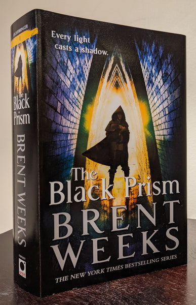 Cover and spine of a hardcover copy of THE BLACK PRISM. A man wearing a hood and cape is standing in a triangular brick tunnel. The man is light from behind; his features are in shadow. The words "Every light casts a shadow" are in the top left corner. The hooded figure is also on the spine, with a yellow stripe at the top and the number one on the stripe.