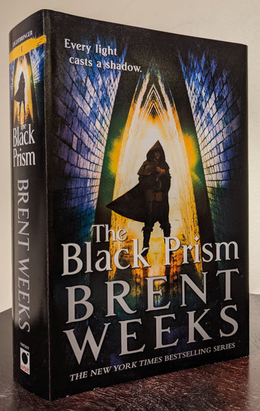 Cover and spine of a hardcover copy of THE BLACK PRISM. A man wearing a hood and cape is standing in a triangular brick tunnel. The man is light from behind; his features are in shadow. The words "Every light casts a shadow" are in the top left corner. The hooded figure is also on the spine, with a yellow stripe at the top and the number one on the stripe.